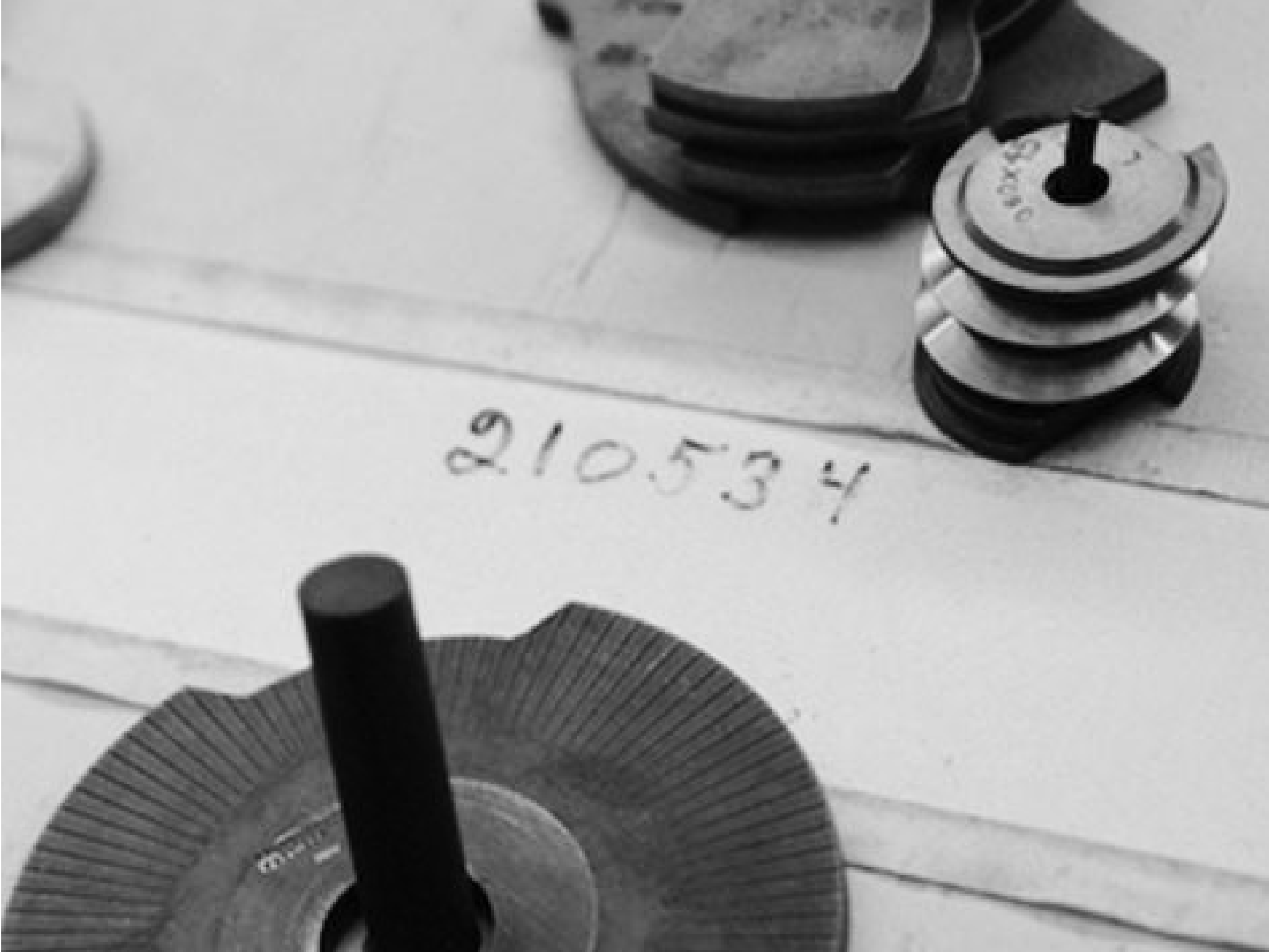 A close-up black and white photo of precision machined components known as cams. The image shows several cams laid out on a surface, with a handwritten number visible in the background. The cams display intricate detailing and precise engineering, showcasing the high level of craftsmanship involved.