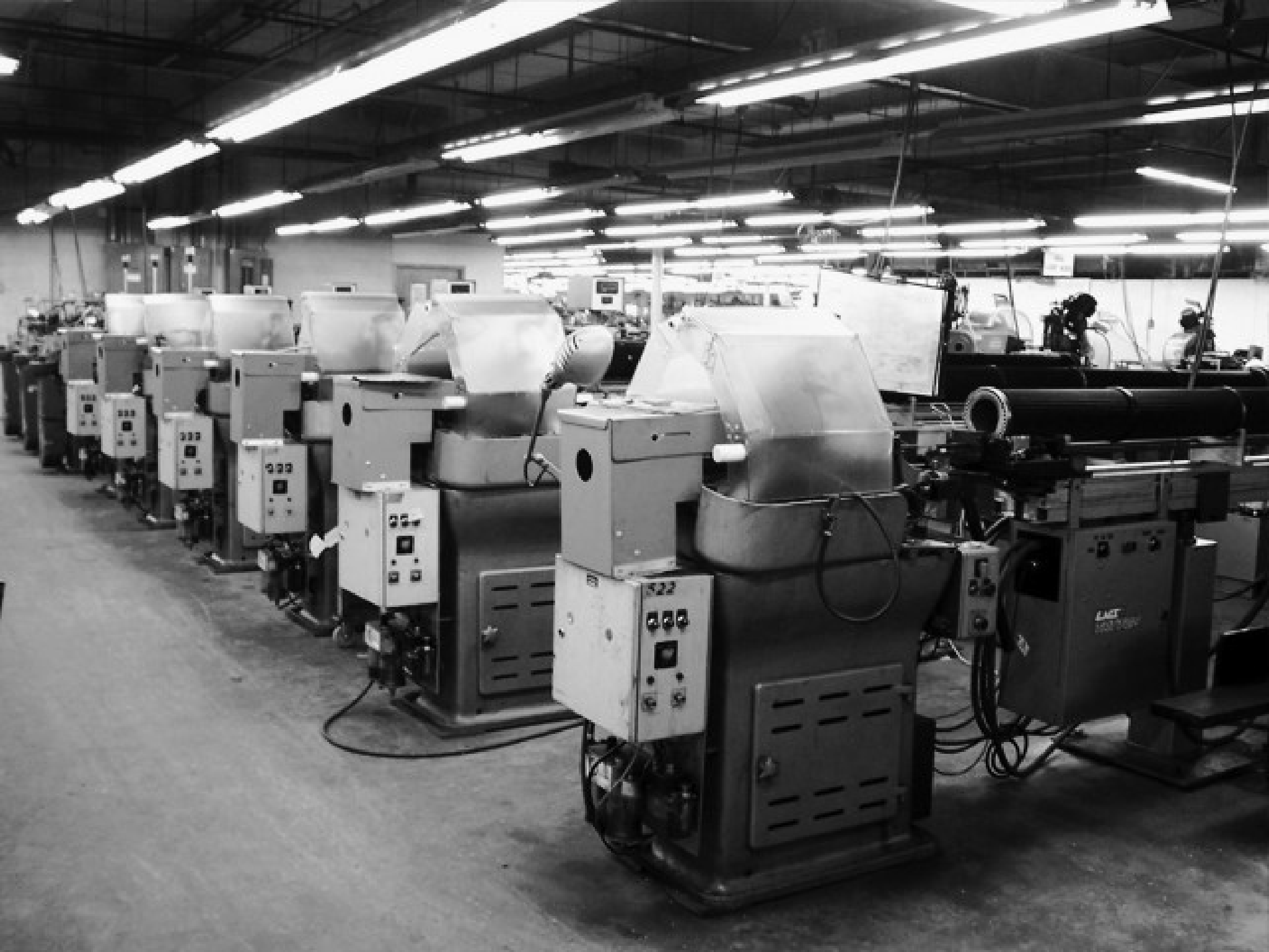 A black and white photo of a row of Escomatic machines in a manufacturing facility. The image shows multiple Escomatic machines lined up, used for precision machining operations. The machines are arranged in a spacious, well-lit environment, highlighting the organized and efficient layout of the workspace.