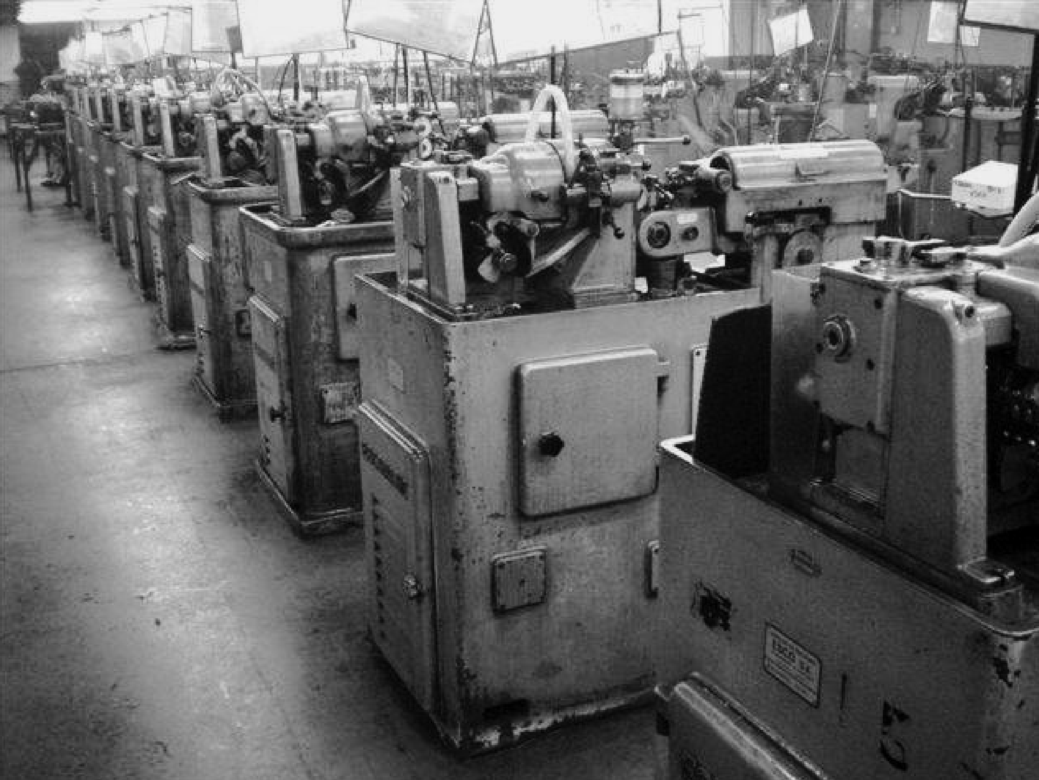 A black and white photo of a row of Escomatic machines in the former location of Iseli Company. The image shows several Escomatic machines lined up, used for precision machining tasks. The machines are sturdy, industrial units, reflecting the organized and efficient setup of the production environment.