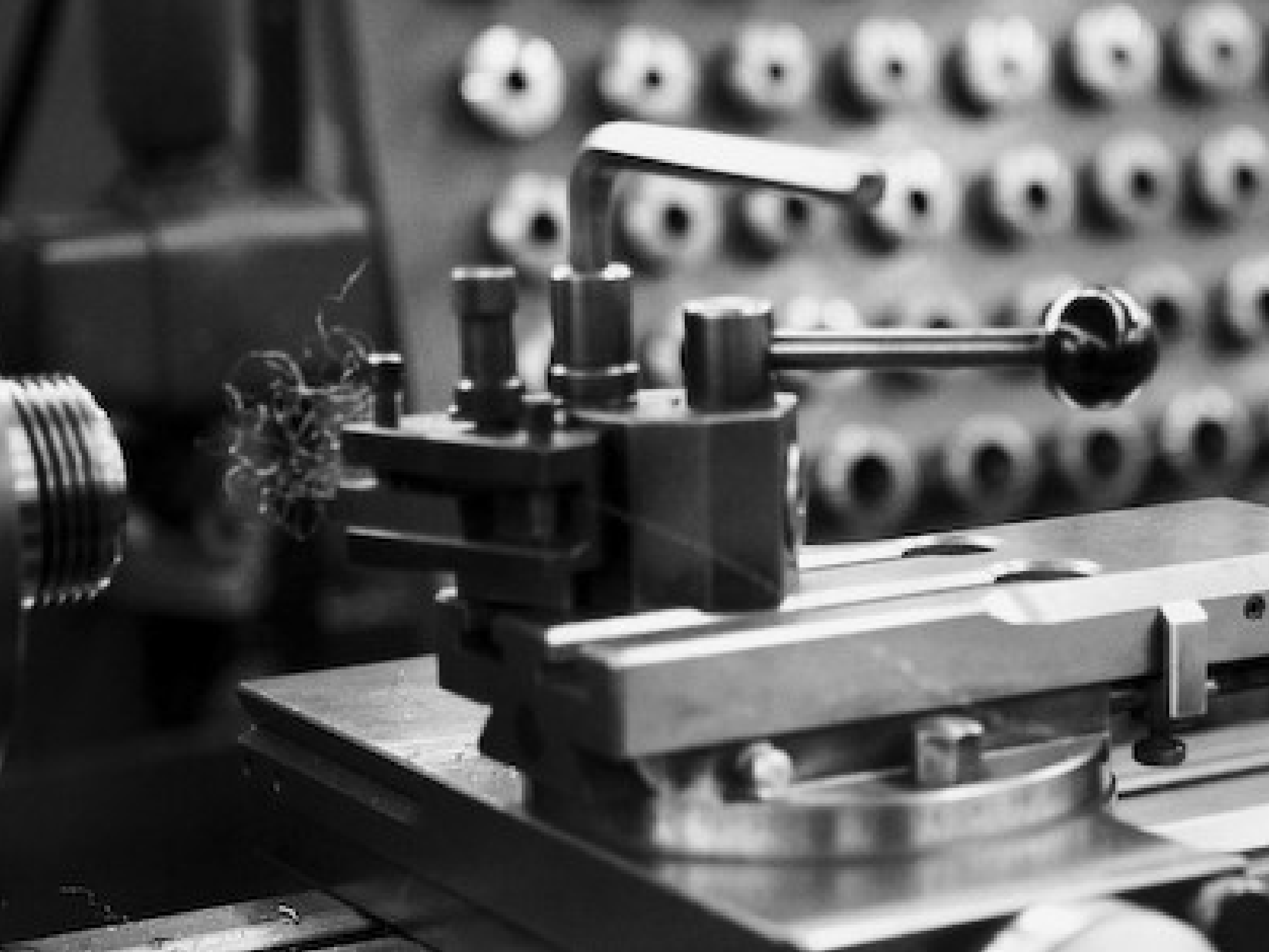 A close-up black and white photo of a precision machining tool in use, capturing the intricate details of the equipment. The image shows metal shavings being produced as the tool operates. The background features an array of parts and tools, highlighting the technical and specialized nature of the machining process.