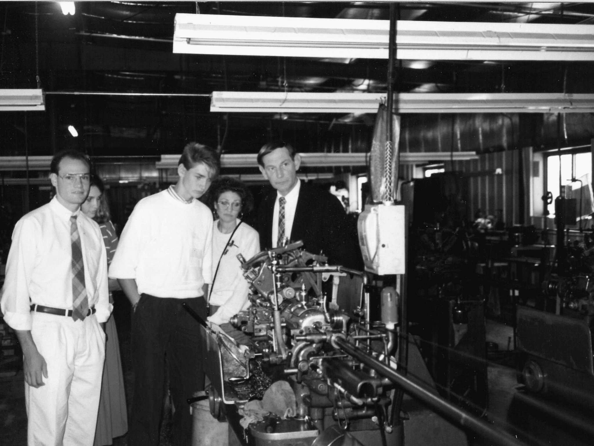 A black and white photo from 1988 showing Mike J. Reader with his father Phil Reader and siblings at Precision Plus's original location in Walworth, WI. The group is inspecting precision machinery in the manufacturing facility, shortly after Phil Reader purchased the business. The setting shows a variety of industrial equipment and tools in the background.