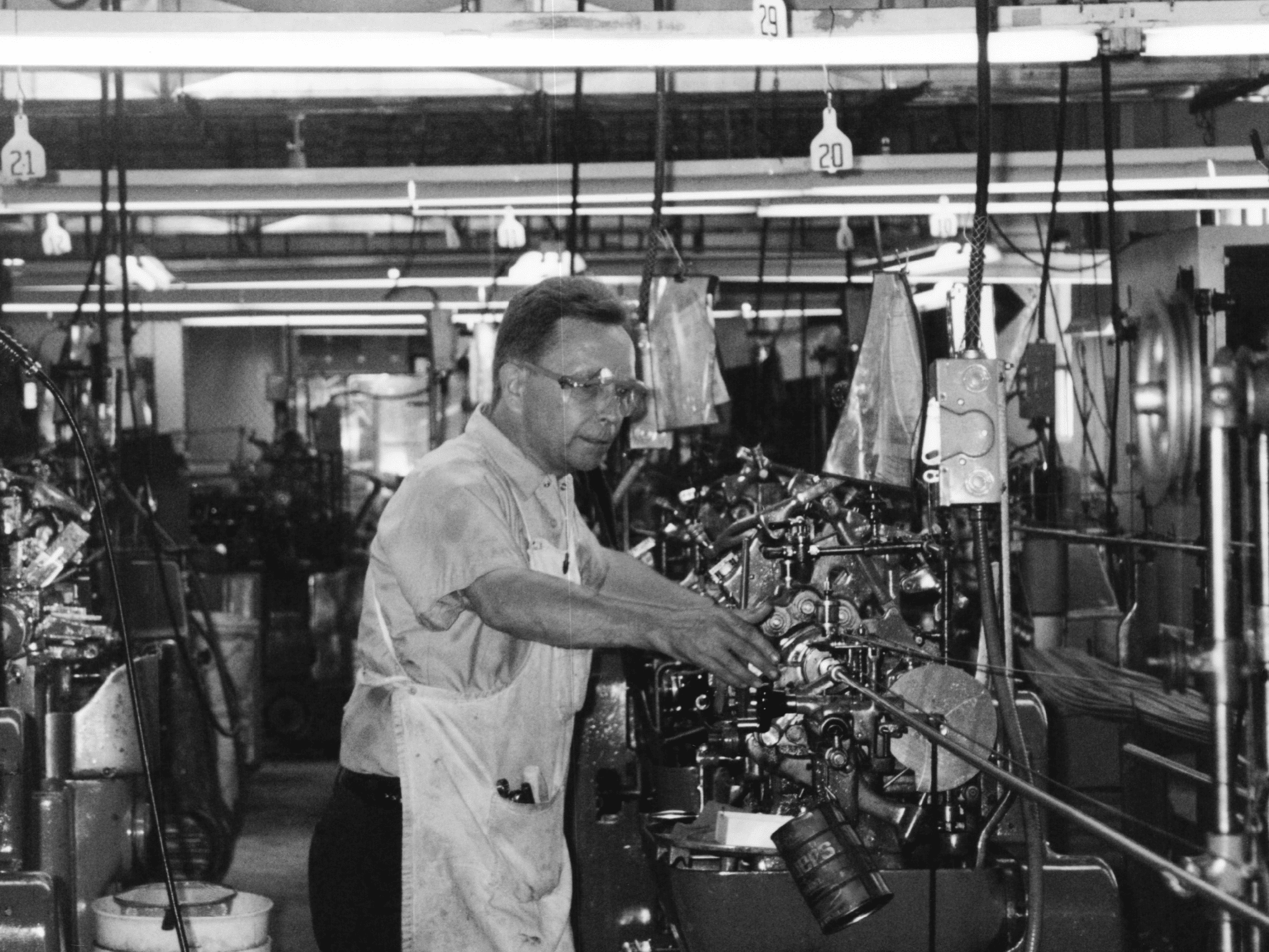 A black and white photo taken in 1998 at the former Precision Plus location in Walworth, WI. The image features a man wearing safety glasses and a work uniform, operating a precision machine. The background displays a row of similar machines and equipment, capturing the industrial setting of the manufacturing facility.